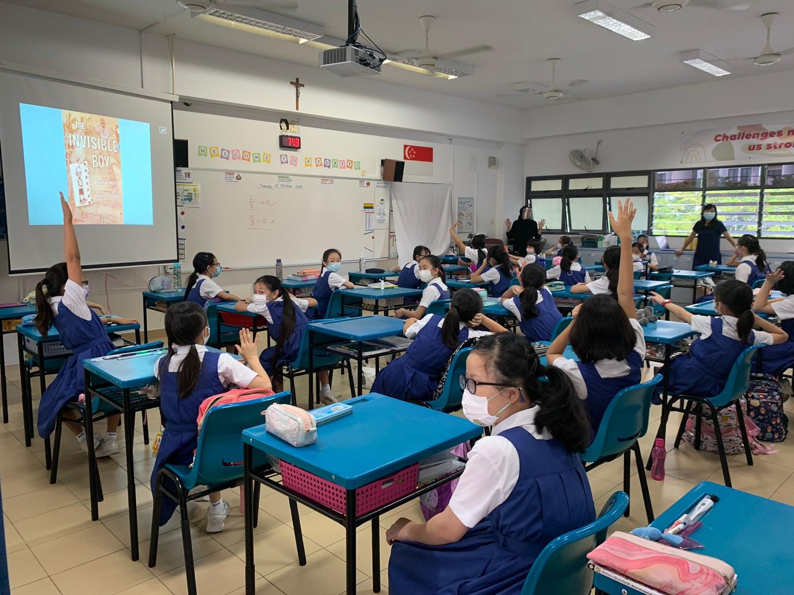 Girls in blue uniforms and masks sit at teal desks, raising hands toward a projected "The Invisible Boy" slide.